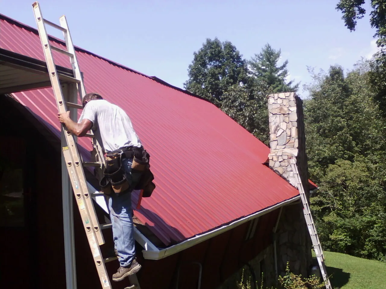 Roofer on a ladder beside a red metal roof and stone chimney — Soffit & Fascia Repair by Roofing Brothers in Fair Oaks